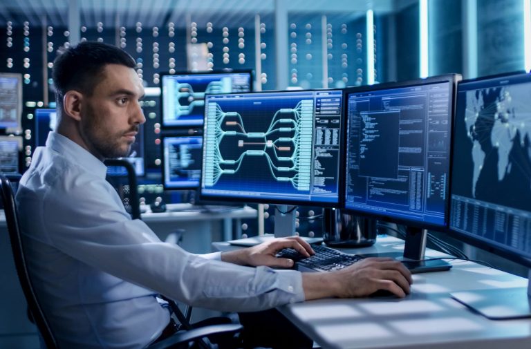 IT professional monitoring network infrastructure and system performance on multiple computer screens in a modern operations center.