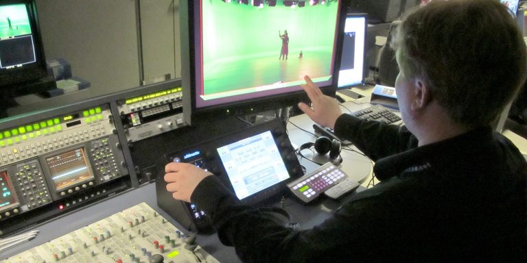 Instructor at Aalto University School using a control panel to demonstrate a technique with a green screen setup in the background.