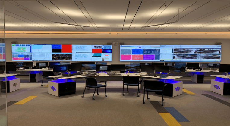 Monitorreihen im Büro einer Leitstelle mit verschiedensten Grafiken und Daten über Versorgung und Transportation abgebildet. Im Vordergrund steht ein Schreibtisch mit drei Stühlen.