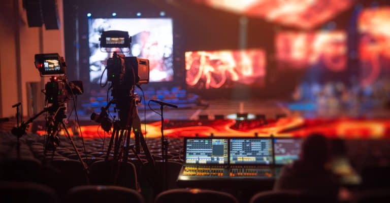 The IHSE broadcast station for the technical crew overlooking an event venue with large screens on a stage, with two cameras and microphones on the left and control panels with small monitors on the right. In front of the display on the far right, the blurry outline of a production staff member is visible.
