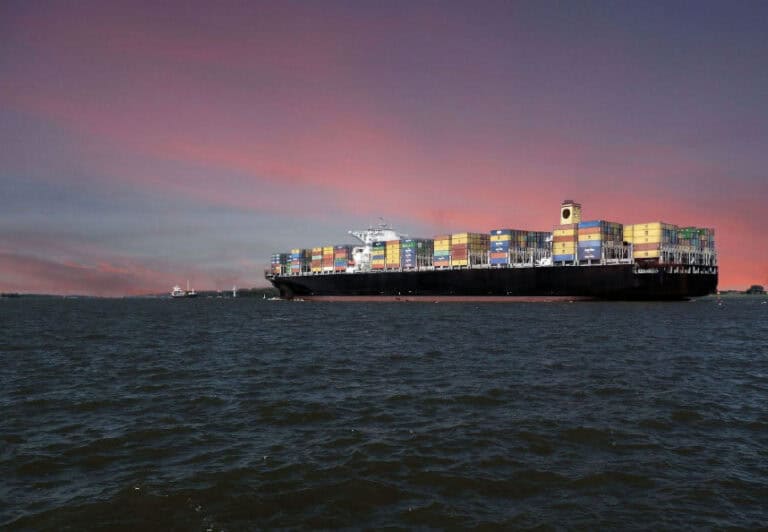 A large cargo ship carrying multicolored shipping containers, sailing on the ocean with a beautiful sunset sky in the background.
